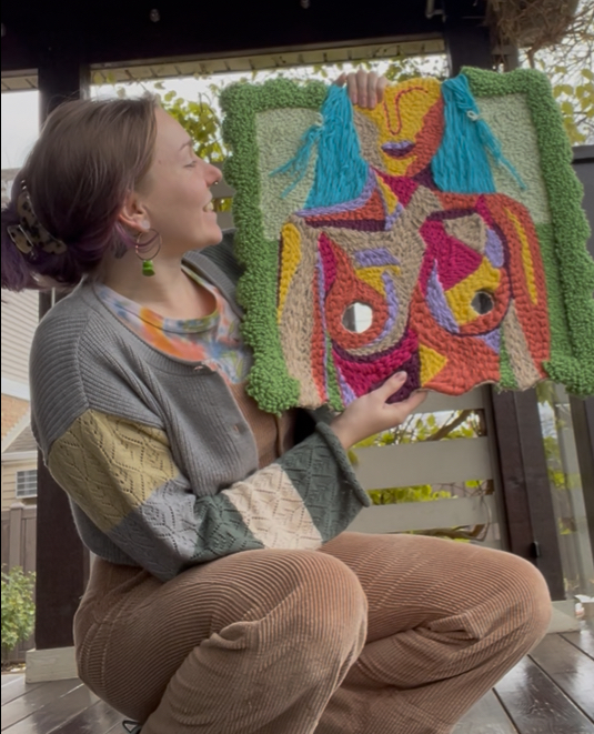 Hope is crouching to the side holding an art piece. The color scheme of the photo includes greens, browns, and colorful pops of pink from the art piece (which is called That's What She Said, and feautured in this sites page Art Dump